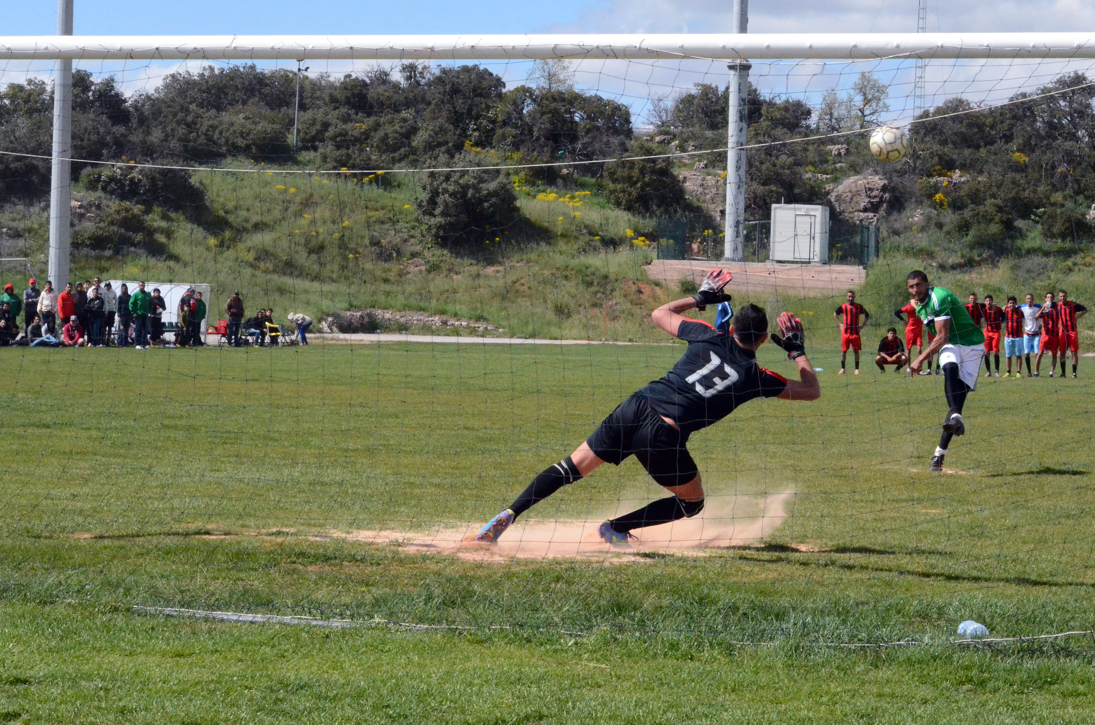 A man in a green jersey takes a penalty during a soccer game which the goalie from the opposite team gets ready to dive towards the ball. The picture is taken from behind the net.